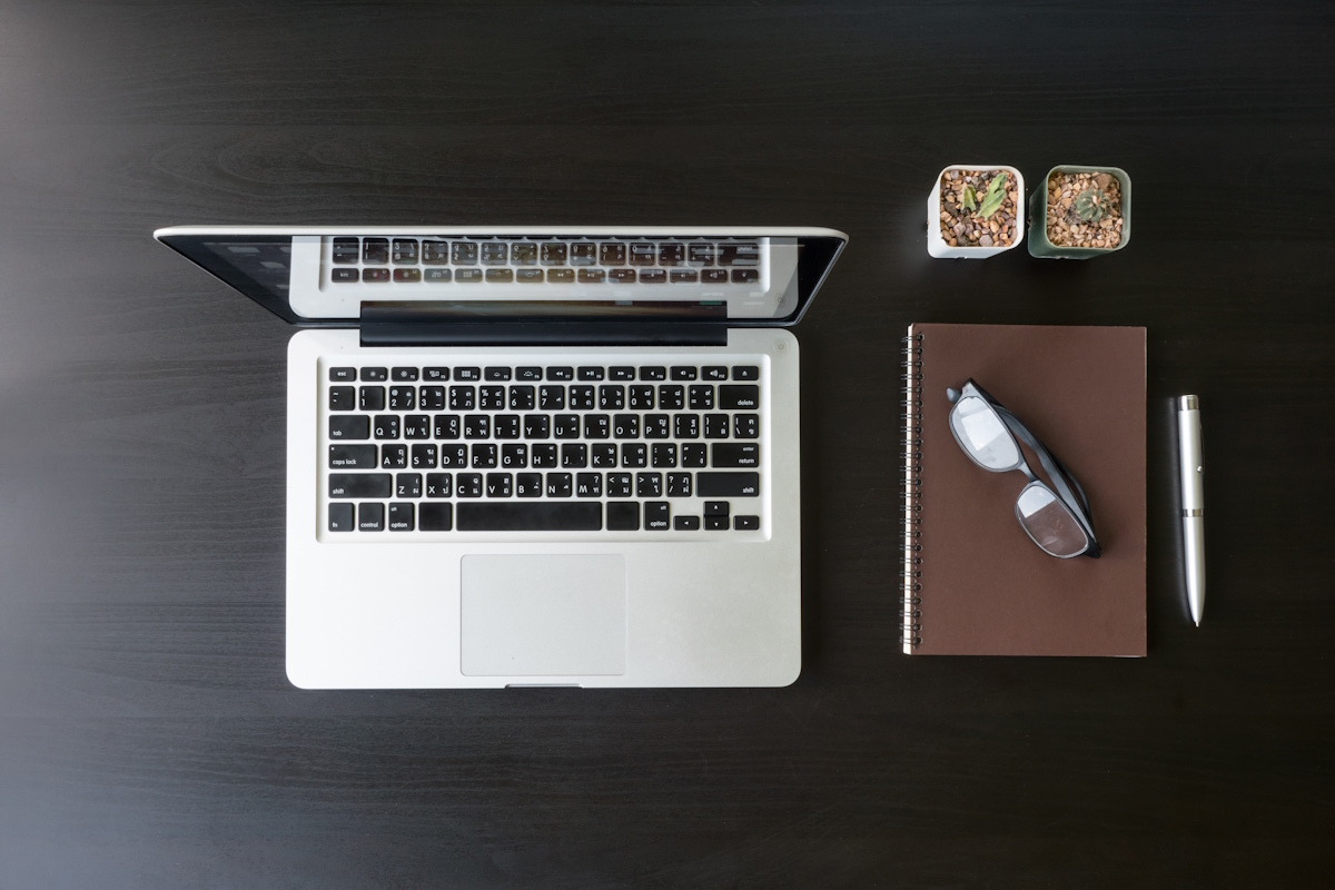 Top view of Laptop with notebook, glasses, pen and cactus on black desk.